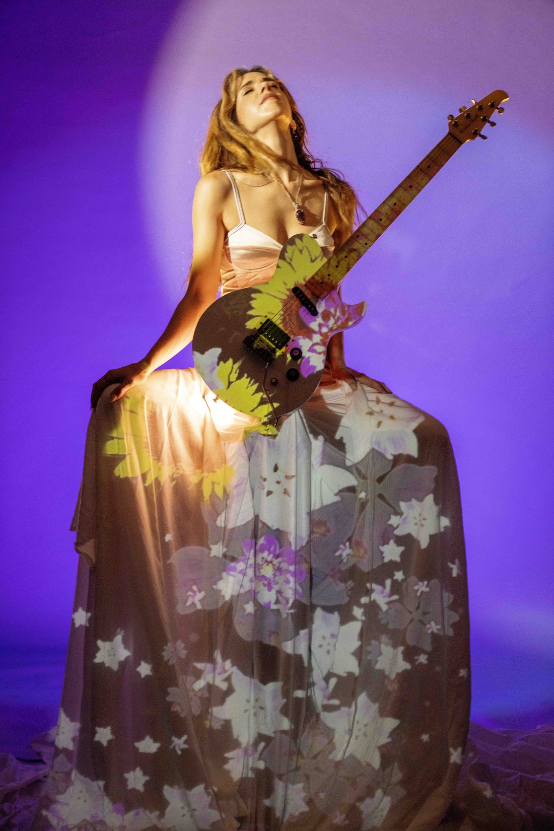 Mar Sánchez con guitarra eléctrica, de pie, entre proyecciones florales amarillas y verdes
